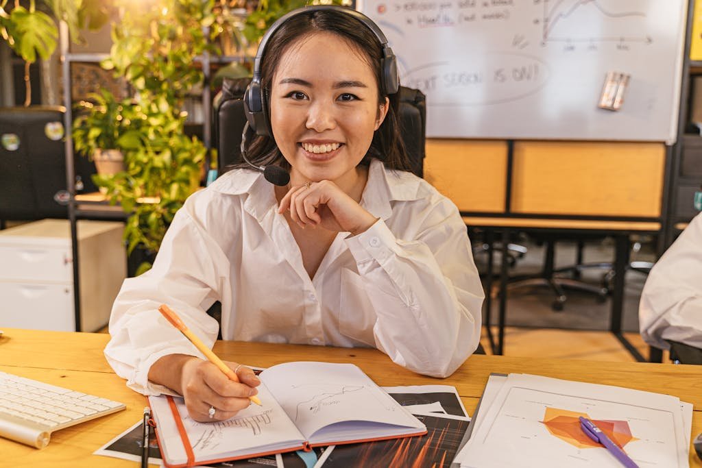A cheerful businesswoman wearing a headset, working on documents in a modern office setting.