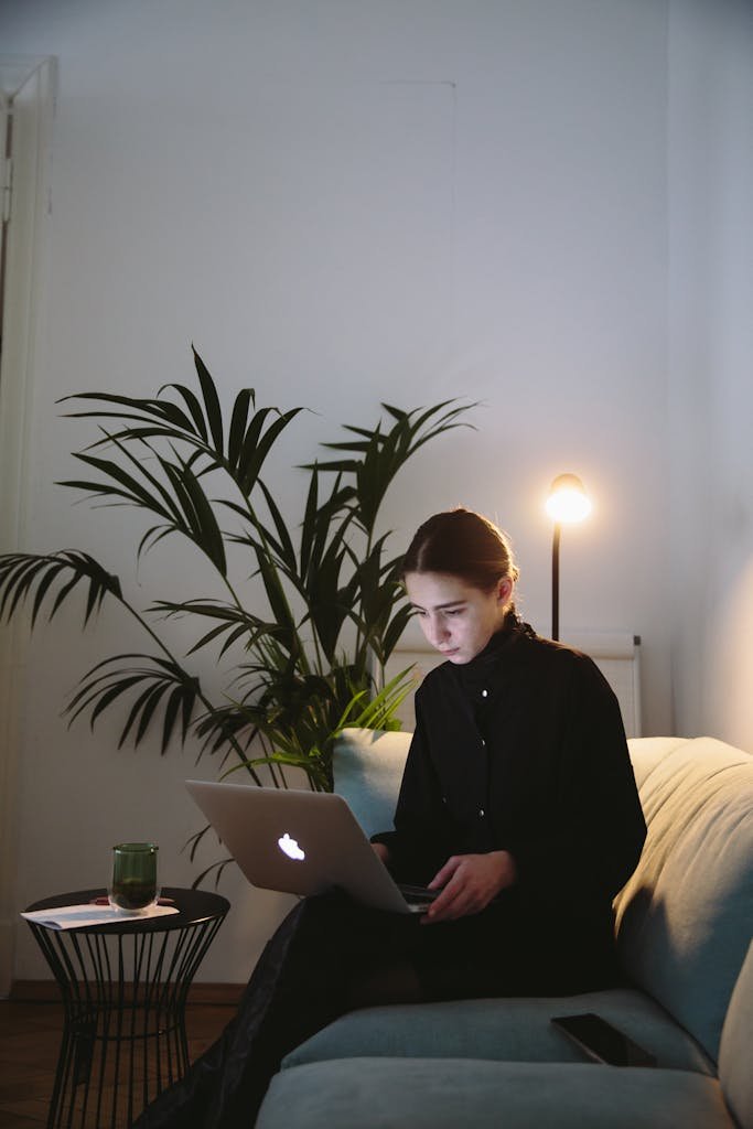 A woman sitting on a sofa working remotely on her laptop with a cozy indoor setup.