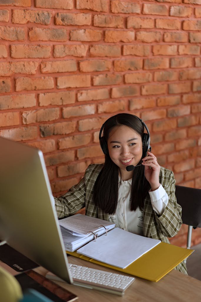 Smiling Asian woman with headset working at computer in office with brick wall background.